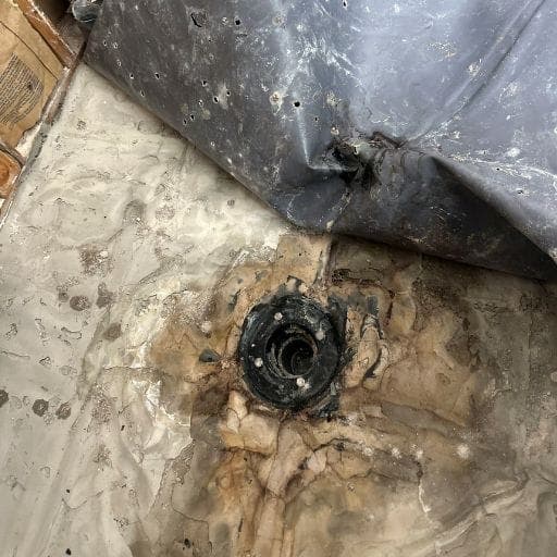 Old shower pan liner peeled back exposing a water-stained mortar bed and drain flange